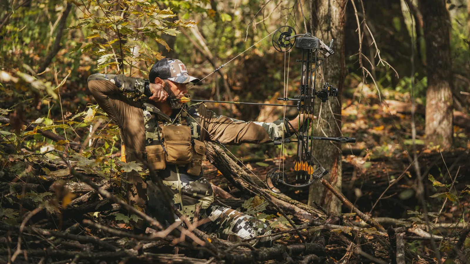 Bowhunter in camo kneeling in thick brush at full draw with a compound bow