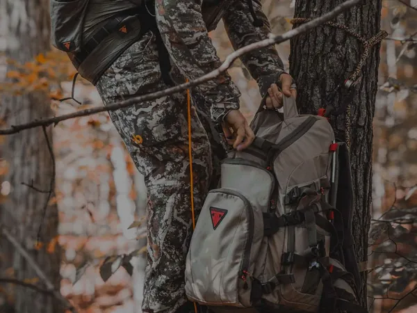 Hunter in a tree saddle reaching into a pack hung on the tree