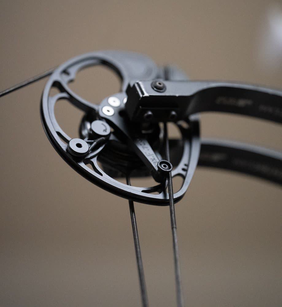 Close-up of a black compound bow cam with its string and cables against a plain beige background.