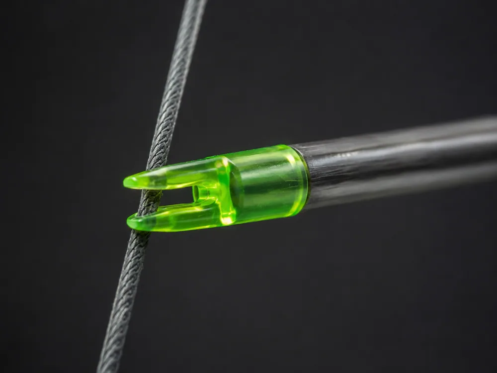 Close-up of a translucent green plastic nock at the rear of a carbon arrow, its slot snapped onto a bowstring against a dark background.